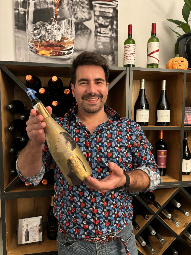 The owner Felipe Risi standing in a wine store, smiling and holding a uniquely designed wine bottle with gold detailing. Behind him, shelves are stocked with various bottles of wine.