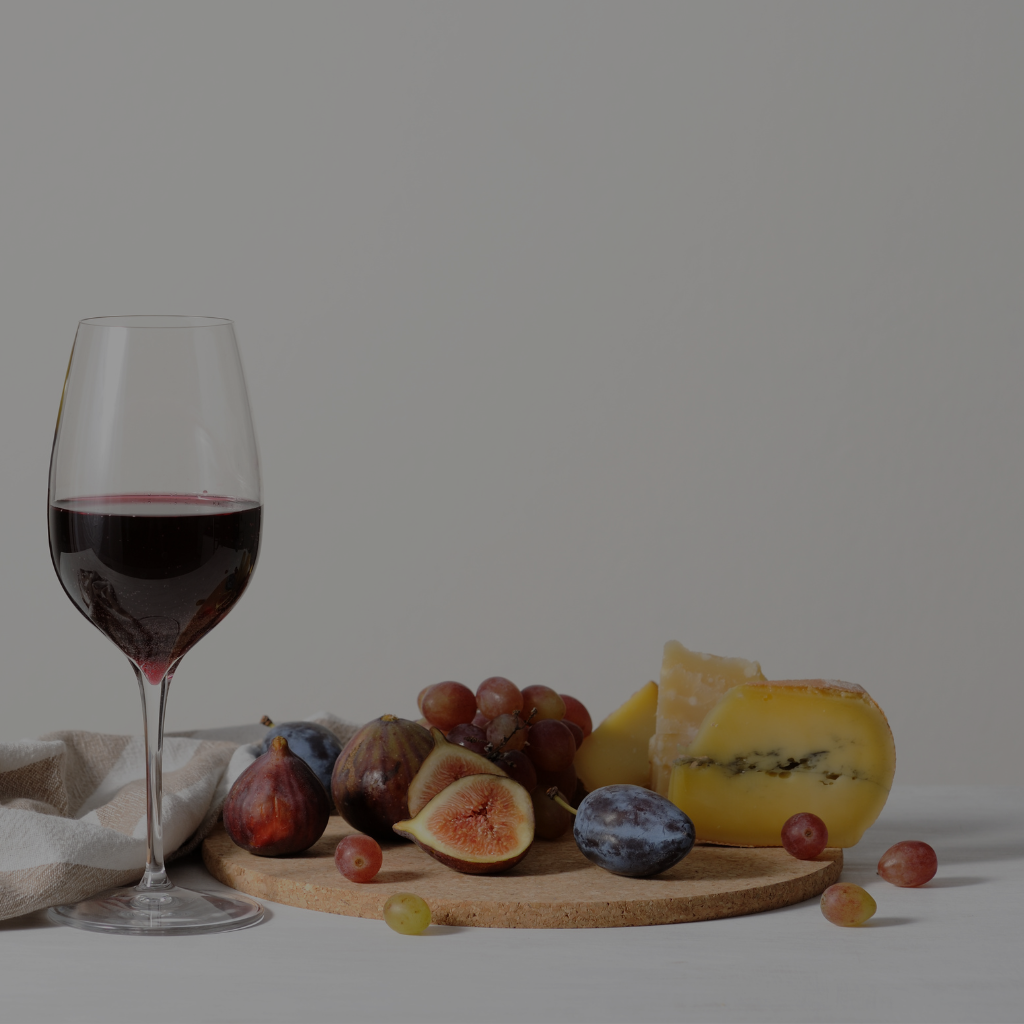 Glass of red wine paired with a wooden platter of assorted cheeses, grapes, figs, and other fruits on a white table with a neutral background.